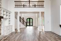Breathtaking two-story foyer features soaring ceilings, elegant wrought iron double doors, stately columns, and a sweeping staircase with iron balusters—anchored by rich wood-look flooring and custom built-ins.