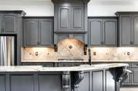 Chef’s kitchen showcases freshly painted solid wood cabinetry, granite countertops, and elegant corbels, all highlighted by a natural stone backsplash and premium stainless appliances.
