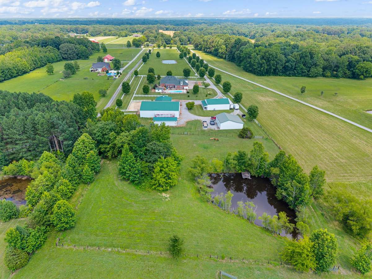 Stunning aerial view showcasing 14.75 acres, including the 3,650 sq ft house, horse barn with indoor riding arena, detached garage with office, 3-bay workshop, pond with dock, and a picturesque, tree-lined driveway.