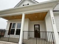 Deep front porch with iron rails and wood ceiling, stained in a natural color and a glass front door.
