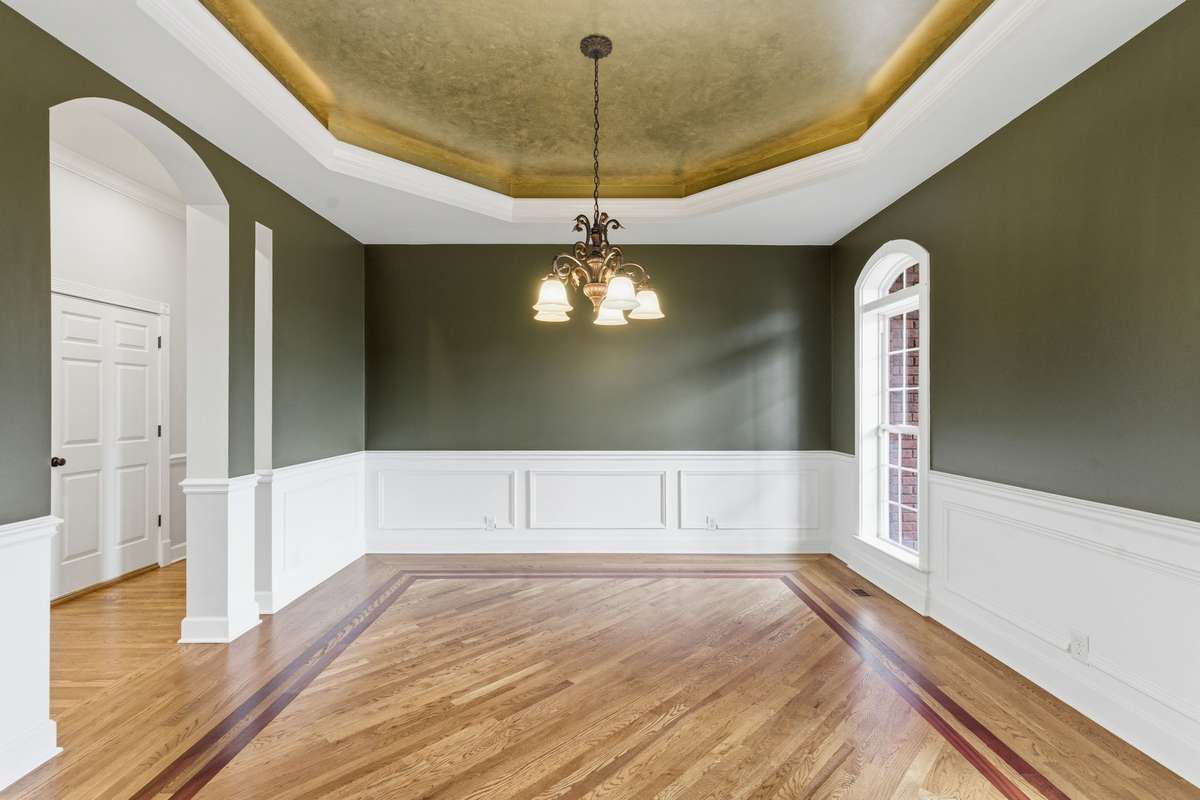 Formal dining room with rich hardwood floors, custom inlay detailing, and elegant wainscoting, all crowned by a dramatic gold tray ceiling and chandelier that create a warm, upscale glow.
