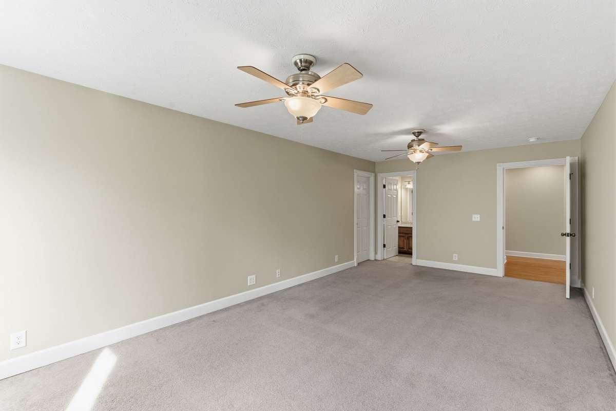 Spacious upstairs secondary primary bedroom featuring two ceiling fans, soft natural light from double windows, a private en-suite bath, and an oversized walk-in closet for exceptional storage.