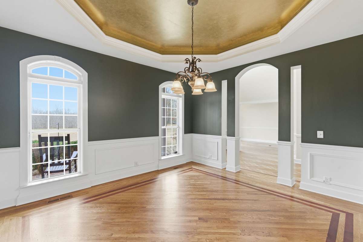 Formal dining room with rich hardwood floors, custom inlay detailing, and elegant wainscoting, all crowned by a dramatic gold tray ceiling and chandelier that create a warm, upscale glow.