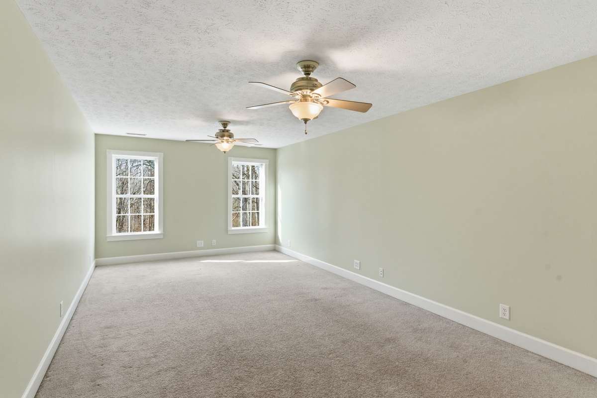 Spacious upstairs secondary primary bedroom featuring two ceiling fans, soft natural light from double windows, a private en-suite bath, and an oversized walk-in closet for exceptional storage.