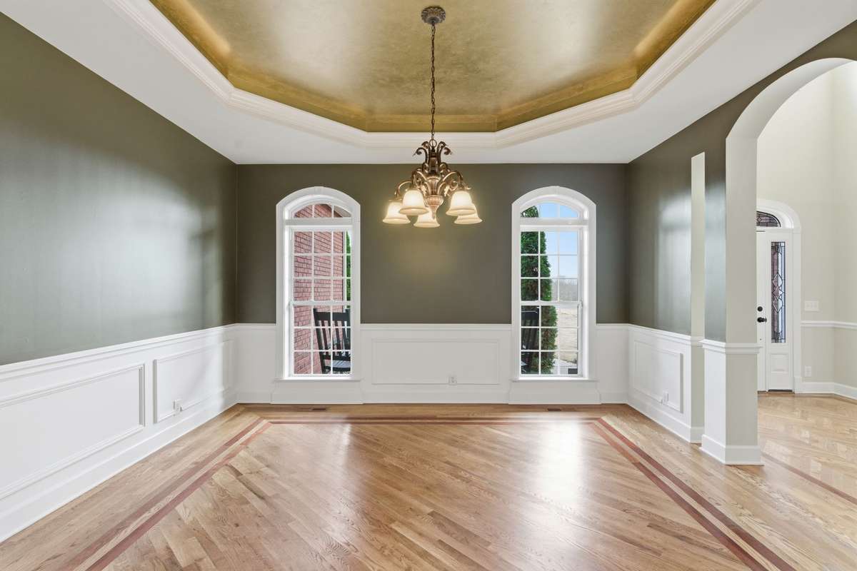 Formal dining room with rich hardwood floors, custom inlay detailing, and elegant wainscoting, all crowned by a dramatic gold tray ceiling and chandelier that create a warm, upscale glow.