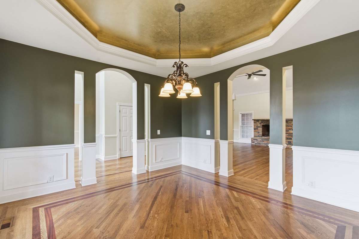 Formal dining room with rich hardwood floors, custom inlay detailing, and elegant wainscoting, all crowned by a dramatic gold tray ceiling and chandelier that create a warm, upscale glow.
