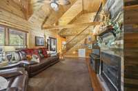 Experience the cozy log home living room featuring warm wood-style flooring, a soaring cathedral ceiling open to the loft, and a captivating double-sided gas log fireplace. Perfect for mountain comfort!