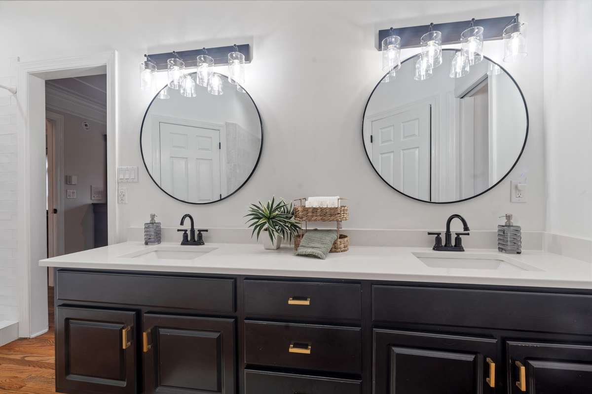 This vanity is a modern double-sink vanity featuring sleek black cabinetry with brass/gold hardware, topped with a clean white countertop and integrated rectangular undermount sinks.