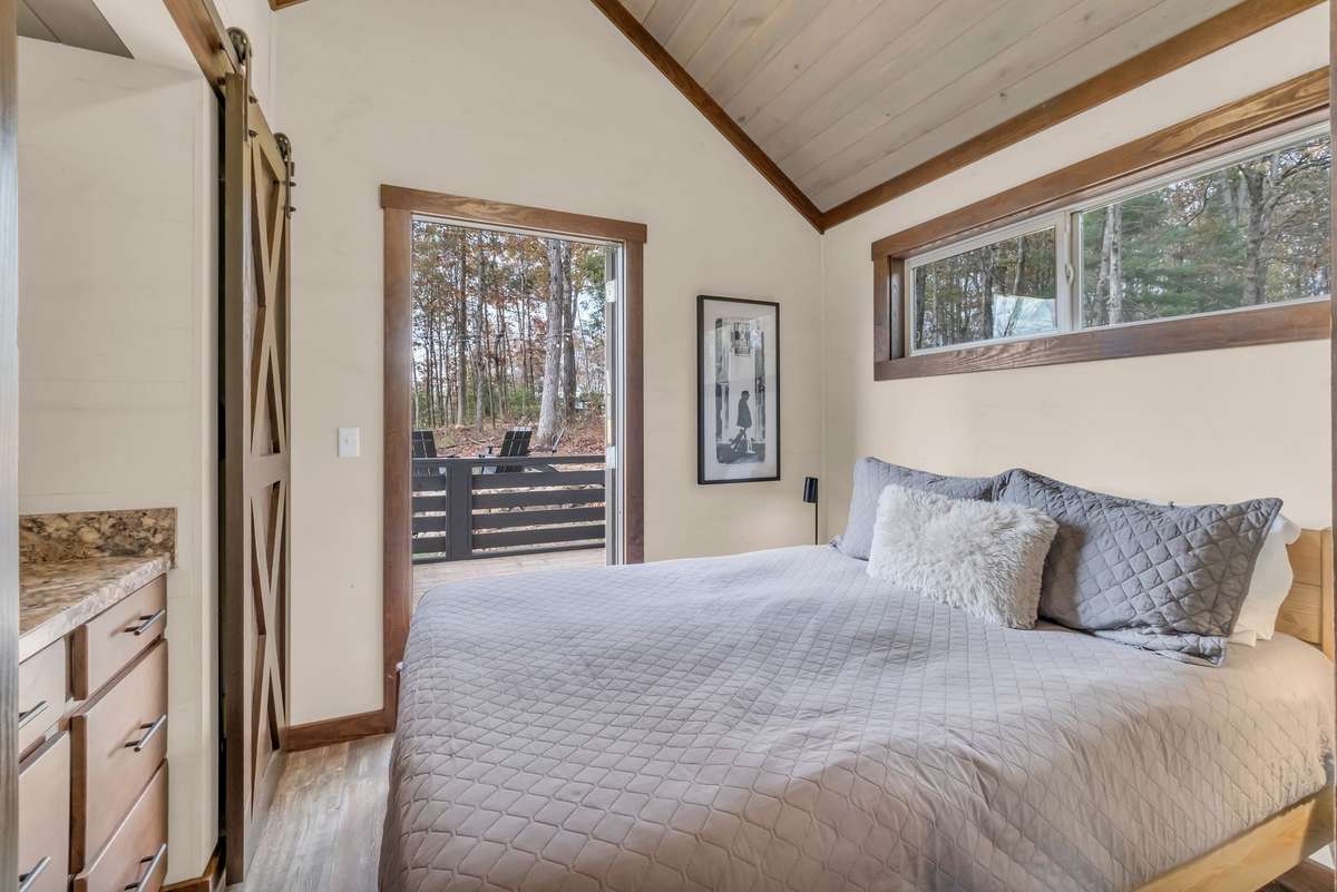 The bedroom has a vaulted ceiling, a large window, a queen size bed and an exterior glass door that opens up to the deck.