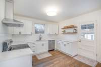 Stainless steel range, dishwasher, and modern vent hood complement the clean look, while a window over the sink brings in natural light. Kitchen has warm hardwood floors, a contemporary flush-mount ceiling light, and stylish floating wood shelves