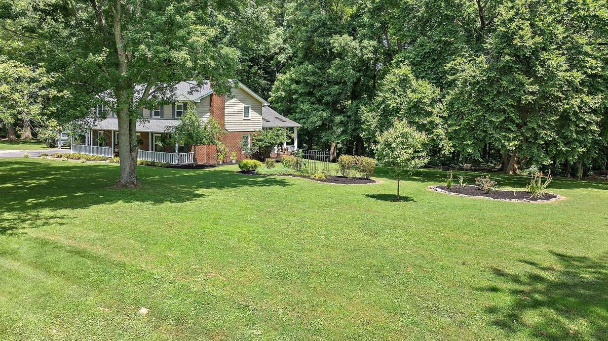 A spacious, well-maintained front yard surrounds the home, complemented by large shade trees and thoughtfully landscaped garden beds. The side view highlights the home’s front and side porches and natural setting.