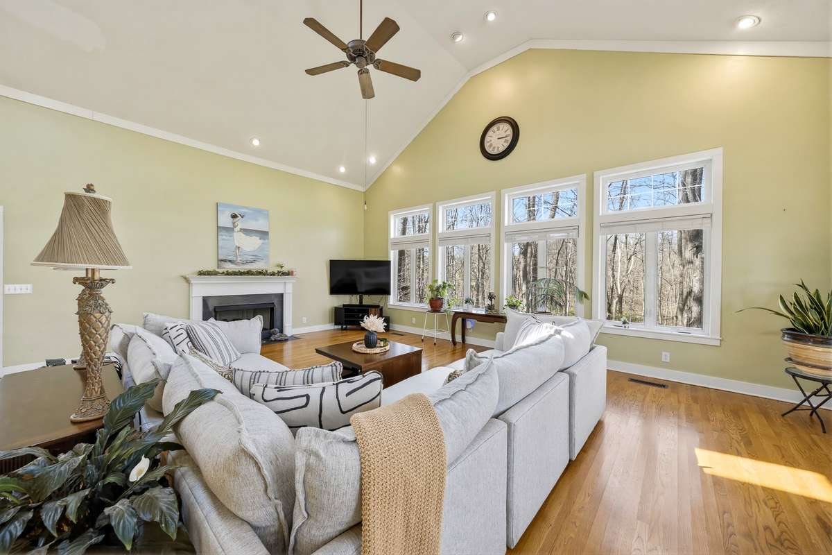 Living Room (Alternate View) – Bright living room with vaulted ceiling, recessed lighting, and a cozy fireplace. Large windows frame scenic views and fill the space with natural light, enhancing the warm hardwood floors.