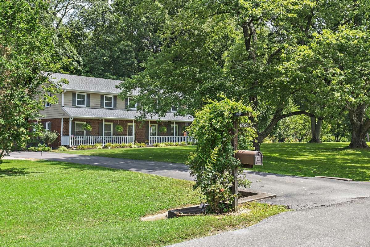 This welcoming front exterior showcases a classic two-story design with a wide front porch, mature landscaping, and a tree-lined setting. The long driveway and charming mailbox garden create an inviting first impression.
