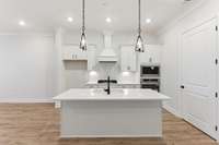 Beautiful kitchen with white cabinetry, gas cooktop, ovens built into the wall cabinet, decorative wood vent hood, island pendents, quartz countertops and tile backsplash to the ceiling.