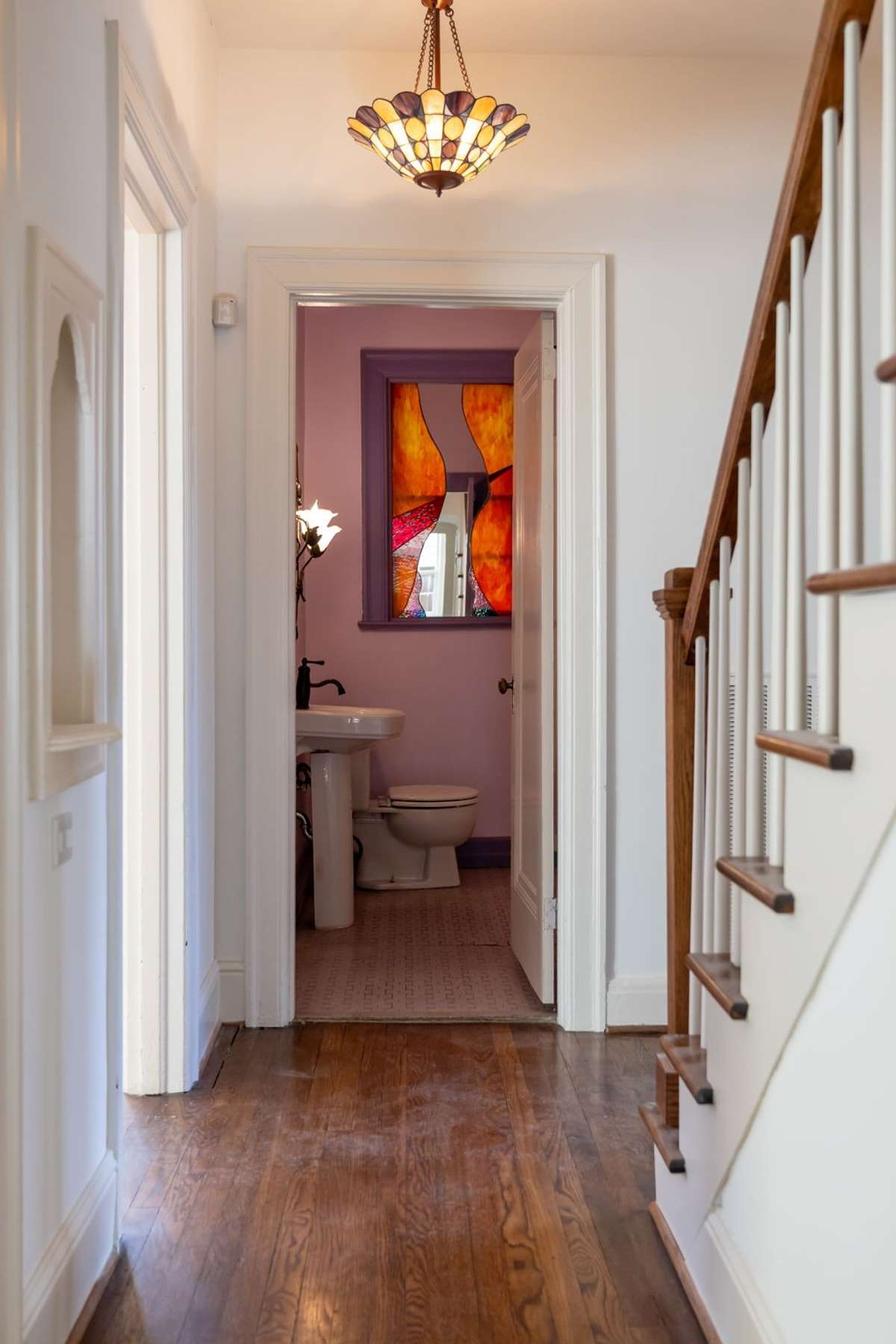 Hardwood floors lead through a bright hallway with period light fixture, wooden staircase detail, and access to the main-level bath.