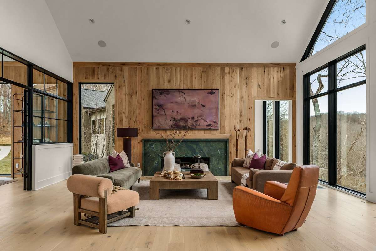 Living room featuring soaring vaulted ceilings, a statement fireplace, and expansive windows that fill the space with natural light while framing peaceful wooded views.