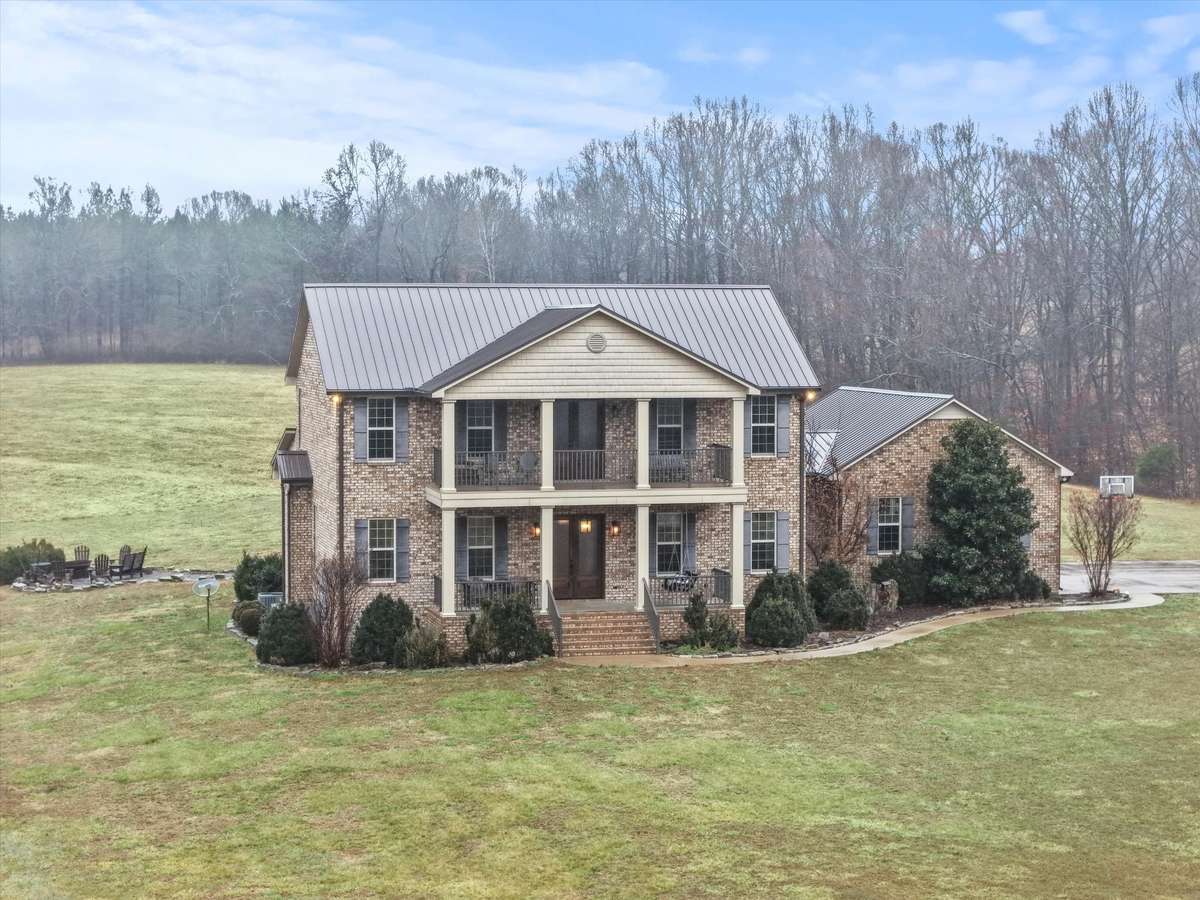 Front of house- Two story full brick house with concrete parking area and sidewalk, private hilltop setting with beautiful views, covered front porch, balcony, classic landscaping, firepit area