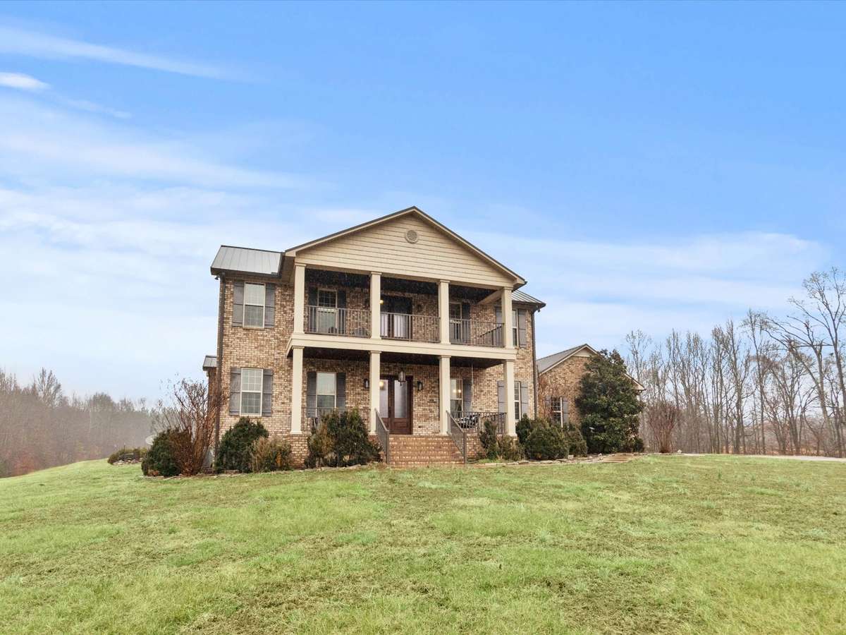 Front of house- Two story full brick house with concrete parking area and sidewalk, private hilltop setting with beautiful views, covered front porch, balcony, classic landscaping, firepit area