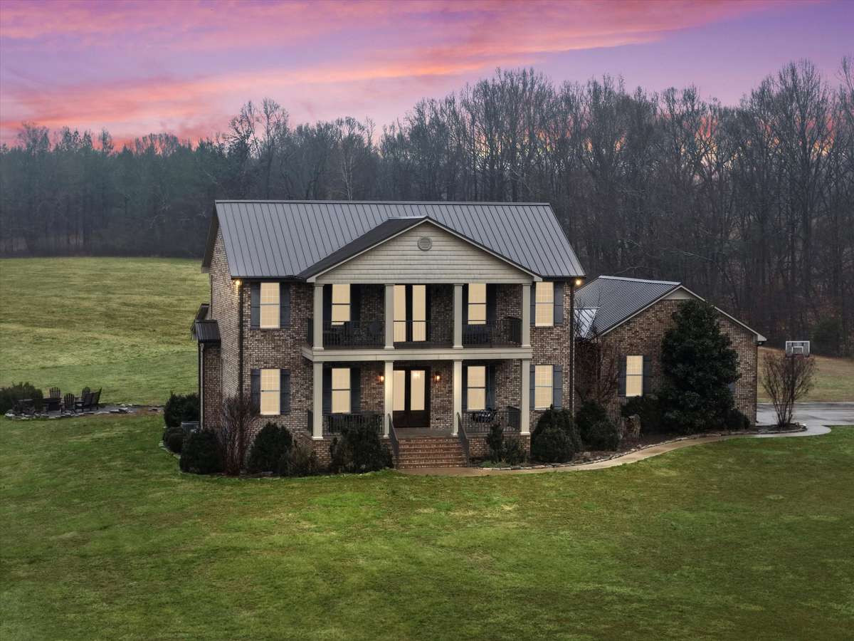 Front of house- Two story full brick house with concrete parking area and sidewalk, private hilltop setting with beautiful views, covered front porch, balcony, classic landscaping, firepit area