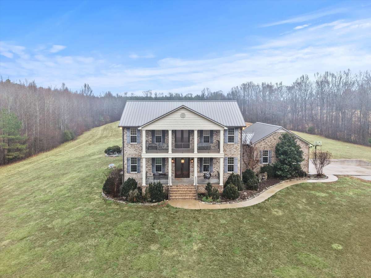 Front of house- Two story full brick house with concrete parking area and sidewalk, private hilltop setting with beautiful views, covered front porch, balcony, classic landscaping, firepit area