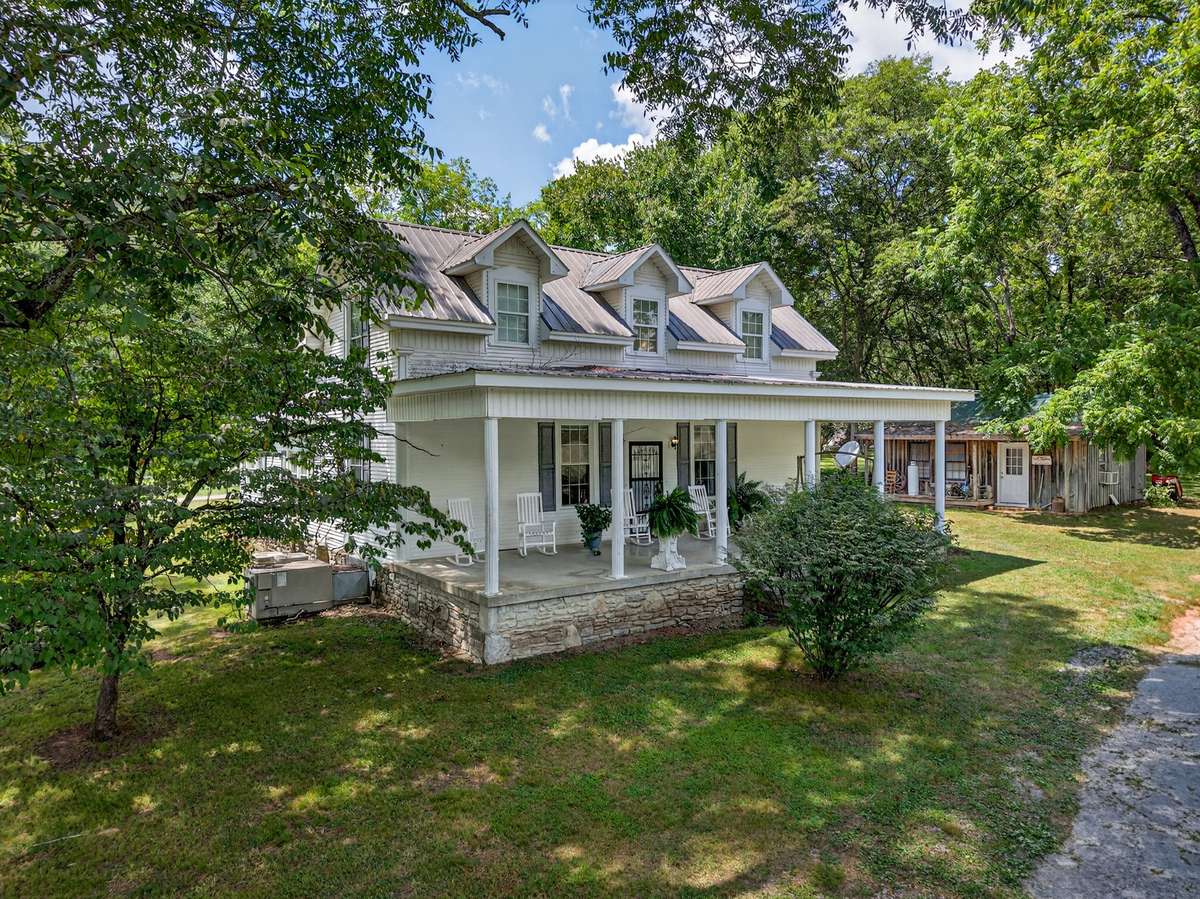 Farmhouse set within a substantial acreage that includes an event center, barn, and supporting structures, highlighted by a covered front porch, metal roof, and wooded surroundings.