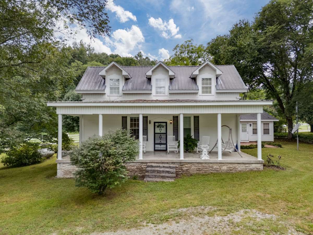 Charming farmhouse with a wide covered front porch, metal roof, and traditional dormers, positioned within expansive acreage that provides privacy, scenic surroundings, and access to the property’s additional structures and event facilities.
