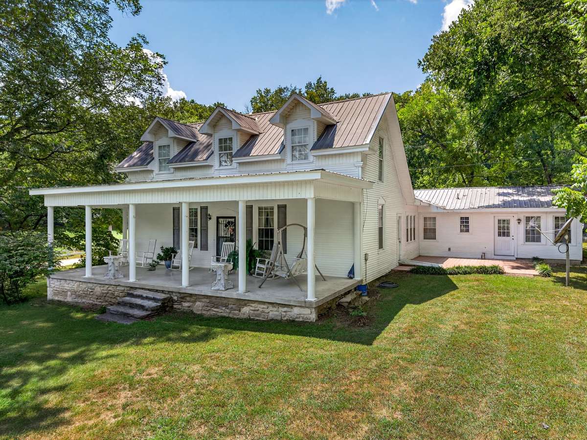 Primary farmhouse located within a large multi-use acreage offering an event pavilion, barn, and open land, complemented by a covered front porch, metal roof, and mature tree setting.
