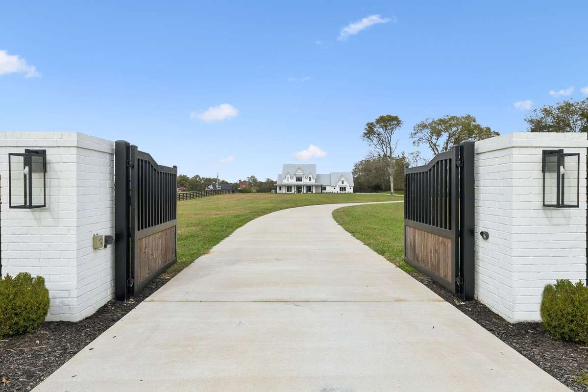 Automatic entry gates open to a long, private drive leading to the residence, creating a secure and welcoming arrival. White brick columns, modern lantern lighting, and wood-accented iron gates frame the approach to this private estate.