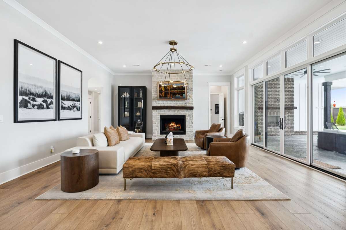 Spacious living room with floor-to-ceiling windows and sliding glass doors opening to the covered patio. Stone fireplace anchors the space, while wide-plank floors and an open layout create an ideal setting for gathering and everyday living.