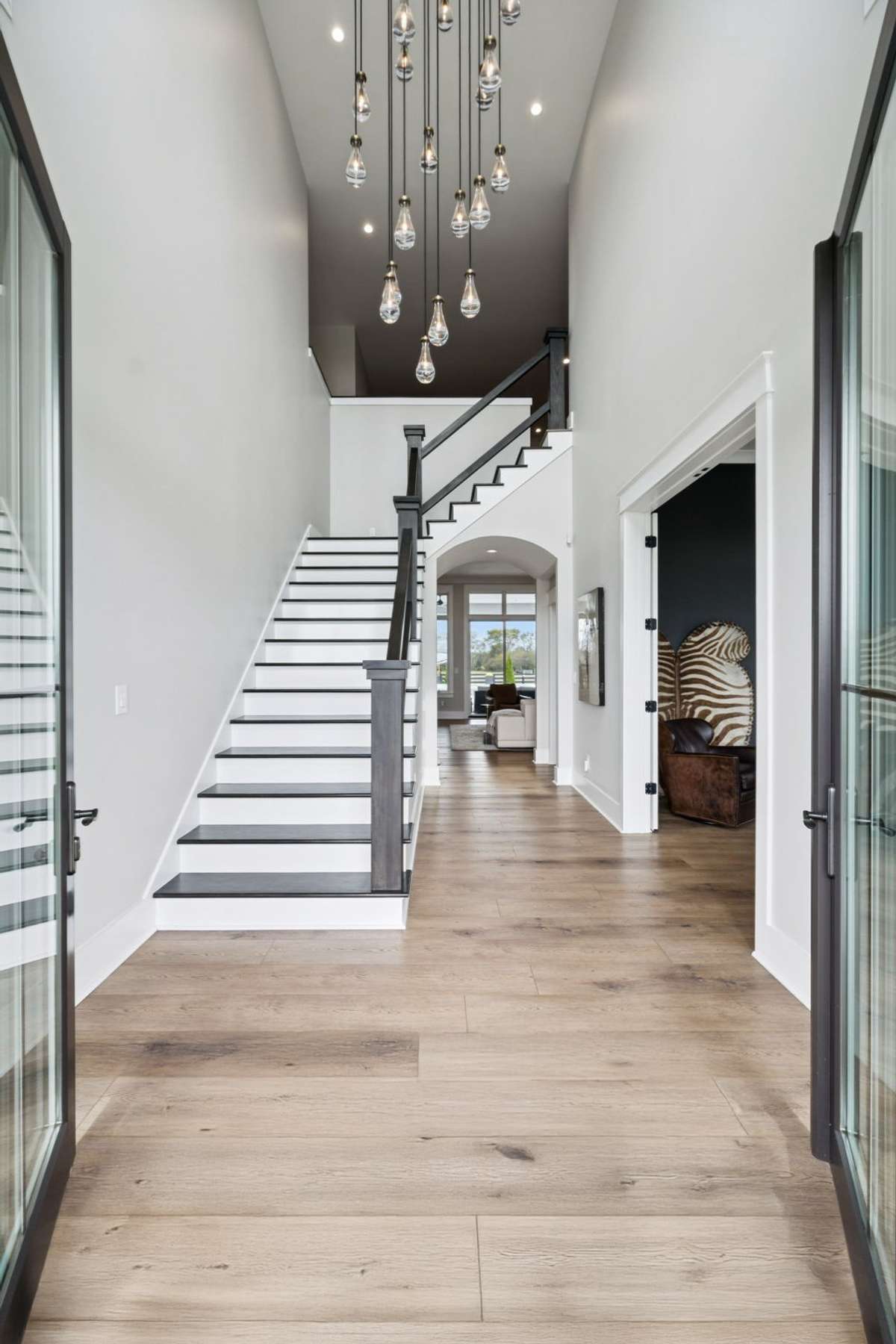 Two-story foyer with soaring ceilings, modern staircase, and statement pendant lighting creates a dramatic welcome. Wide-plank floors lead into the main living areas, offering an open layout and seamless flow throughout the home.