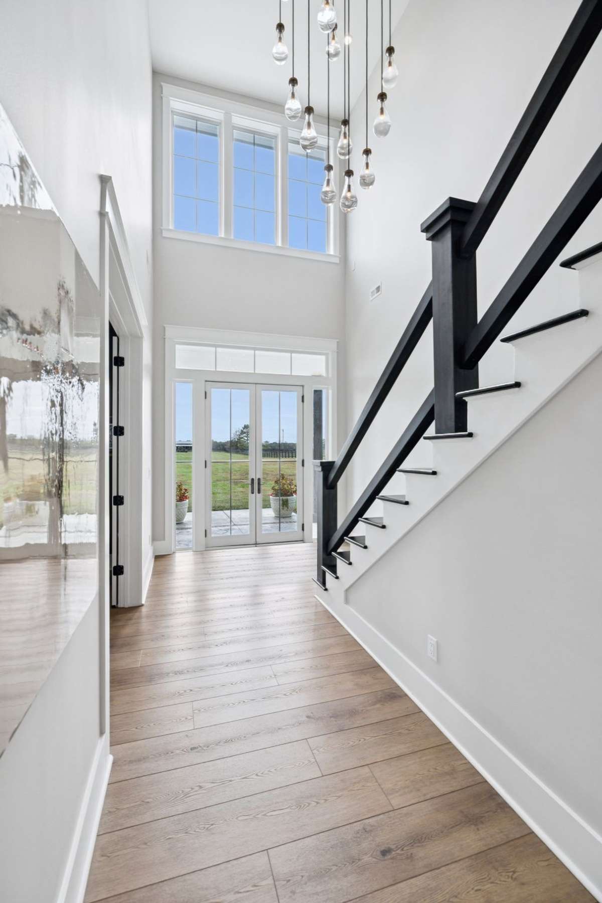 Light-filled entry with floor-to-ceiling windows and glass doors framing views of the acreage beyond. Modern staircase with contrasting railing detail and wide-plank floors create a clean, refined transition into the heart of the home.