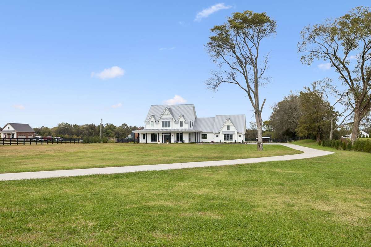 Sweeping concrete drive curves through open, level acreage, creating a picturesque approach to the home. Expansive lawn offers room for recreation, animals, or future additions, all framed by black fencing and mature trees for added privacy.