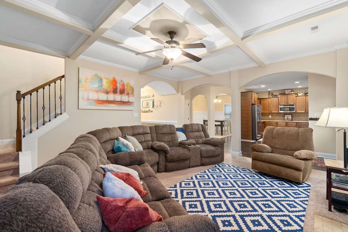 Expansive living room with a coffered ceiling, ceiling fan, and open sightlines to the kitchen and staircase, offering a bright and inviting space for relaxing and entertaining.
