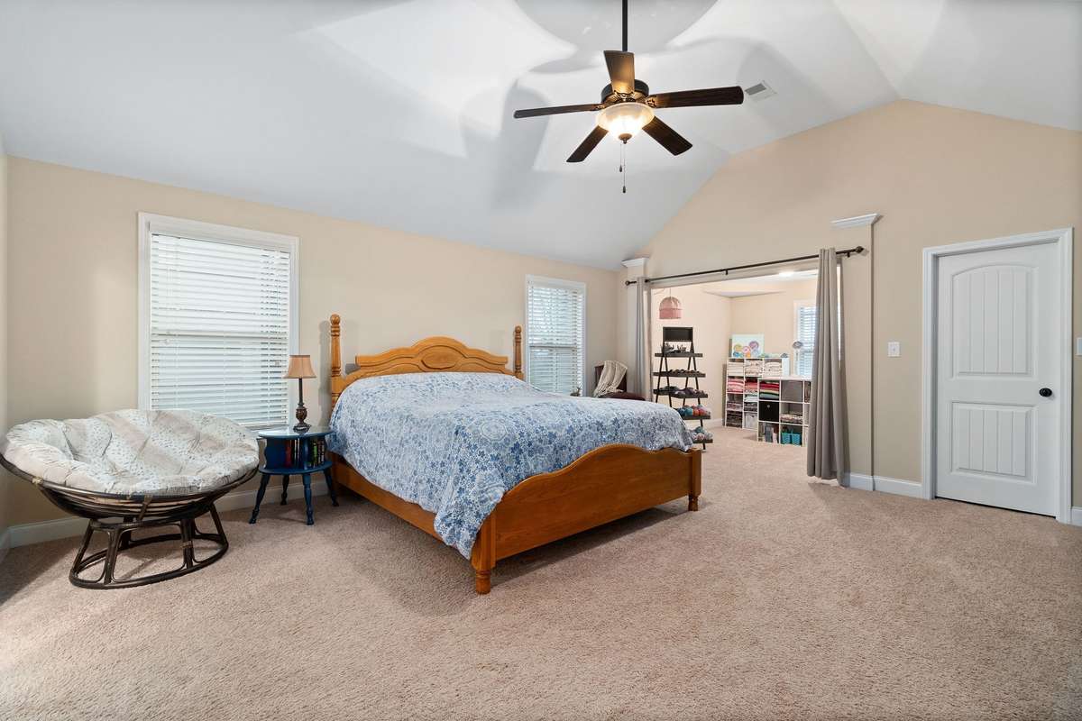 Spacious primary suite featuring vaulted ceilings, ceiling fan, plush new carpet, dual windows for natural light, and a peek into the private sitting nook or walk-in closet area beyond.