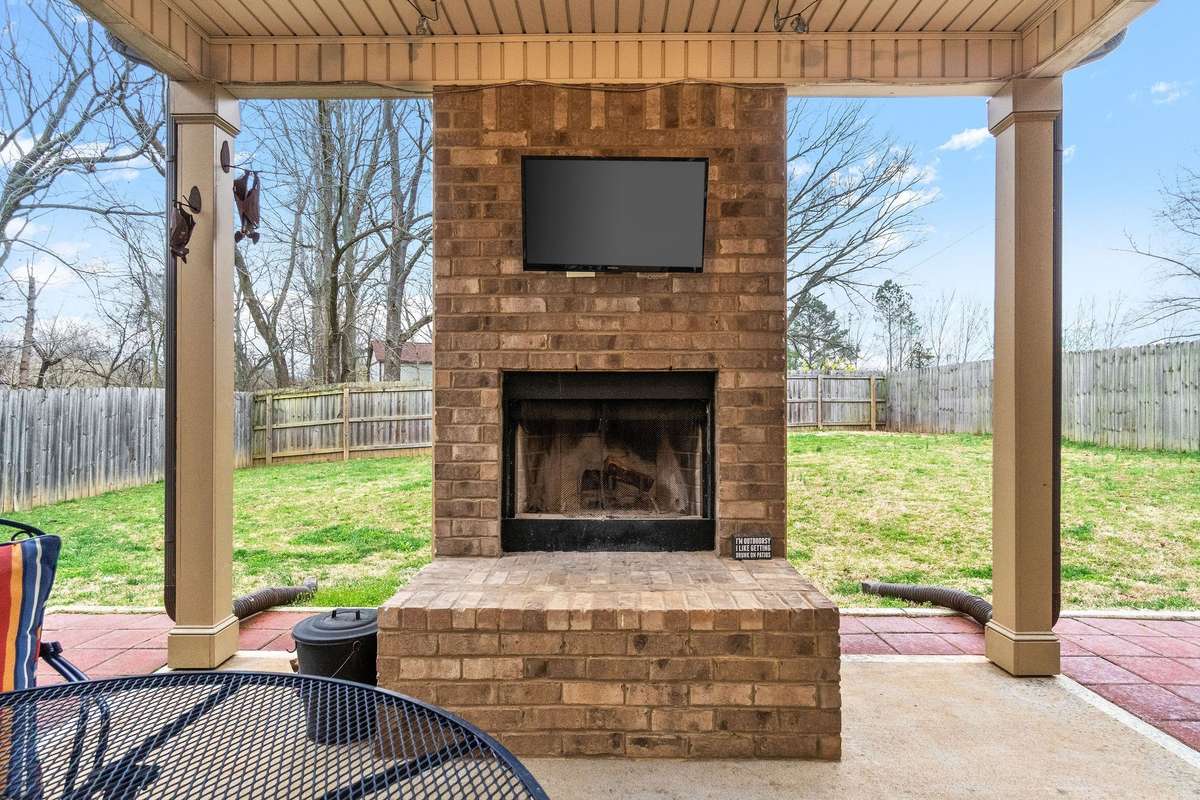 Close-up of the stunning outdoor brick fireplace with mounted TV under the covered back deck — the ultimate centerpiece for outdoor entertaining within the privacy-fenced backyard.