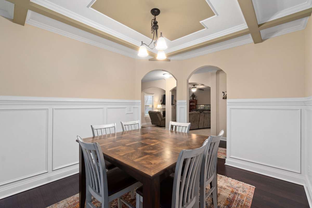 Elegant dining area featuring a dramatic coffered ceiling with chandelier, detailed wainscoting, rich hardwood floors, and graceful arched doorways connecting to the main living spaces.