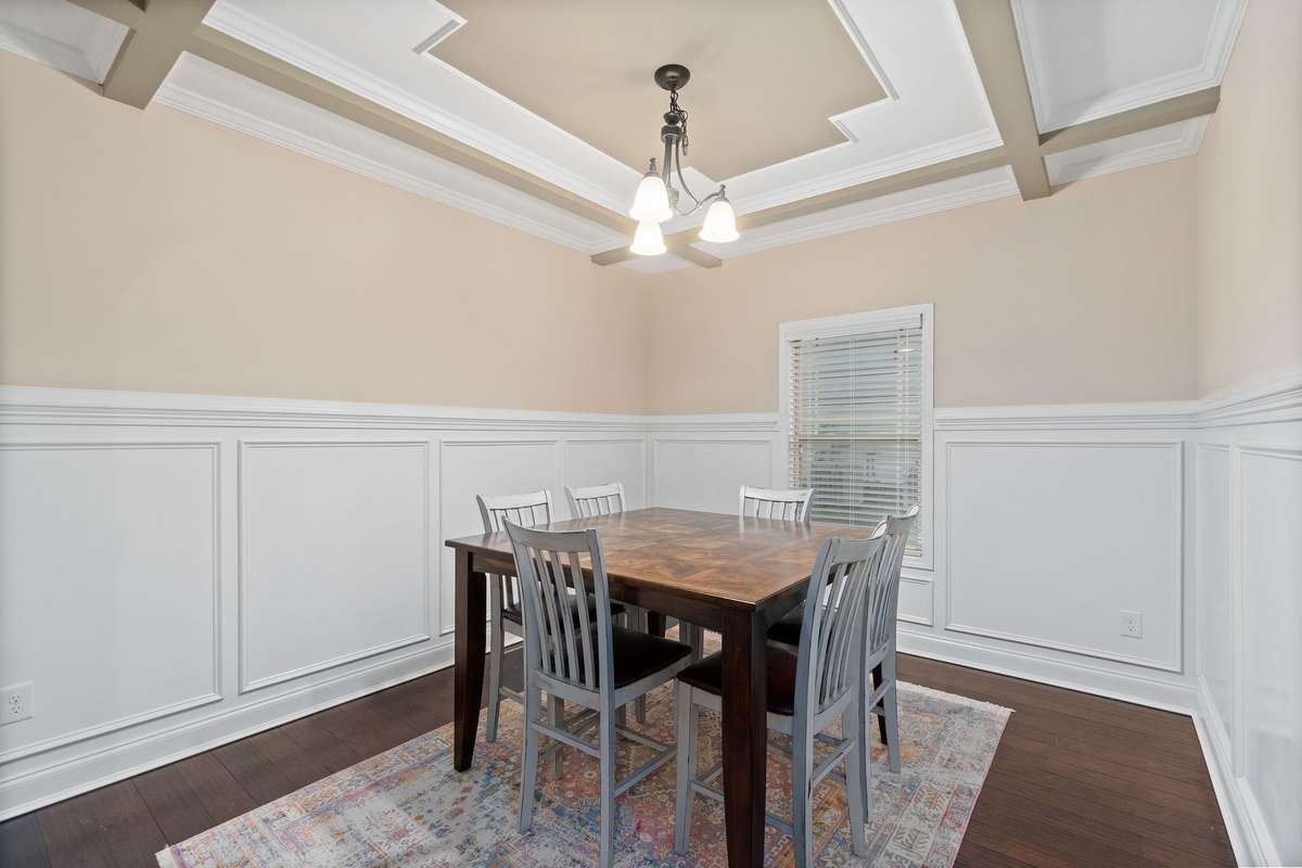 Formal dining room with an impressive coffered ceiling, designer chandelier, elegant board-and-batten wainscoting on all walls, and gleaming hardwood floors — ideal for hosting gatherings.