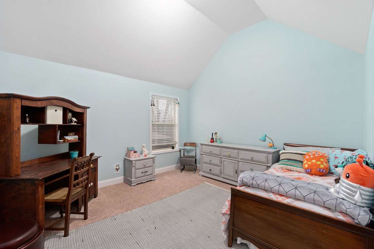 Comfortable secondary bedroom with soft blue-green accent walls, vaulted ceiling, fresh carpet, and a window providing natural light, great for kids, guests, or a home office setup.