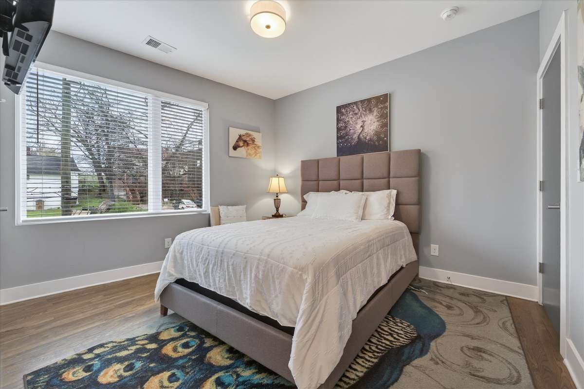 Bedroom 1 offers a serene, stylish retreat—highlighted by a chic peacock rug, tasteful wall decor, and a bright window, creating a restful, inviting atmosphere in this fully furnished suite.