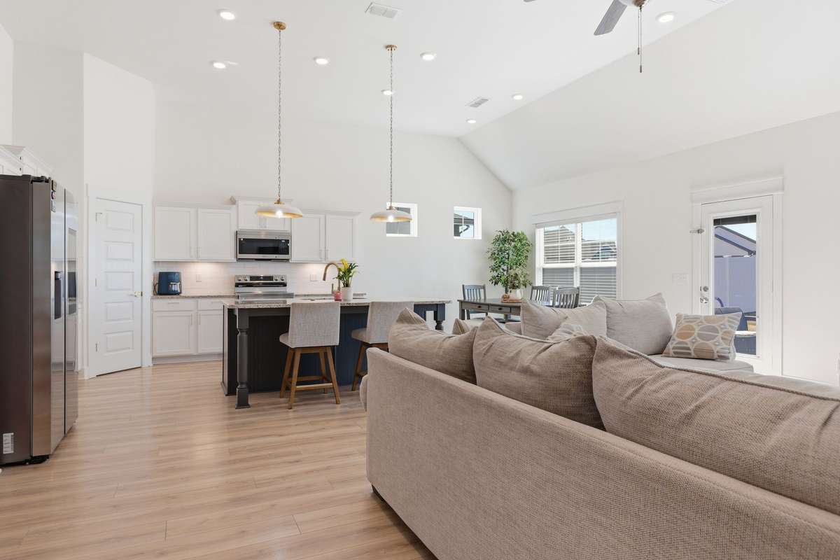 Seamless flow between the kitchen, dining, and living spaces. The kitchen offers white cabinetry, stainless steel appliances, and a large island with seating—perfect for entertaining or everyday living. Natural light pours in through multiple windows