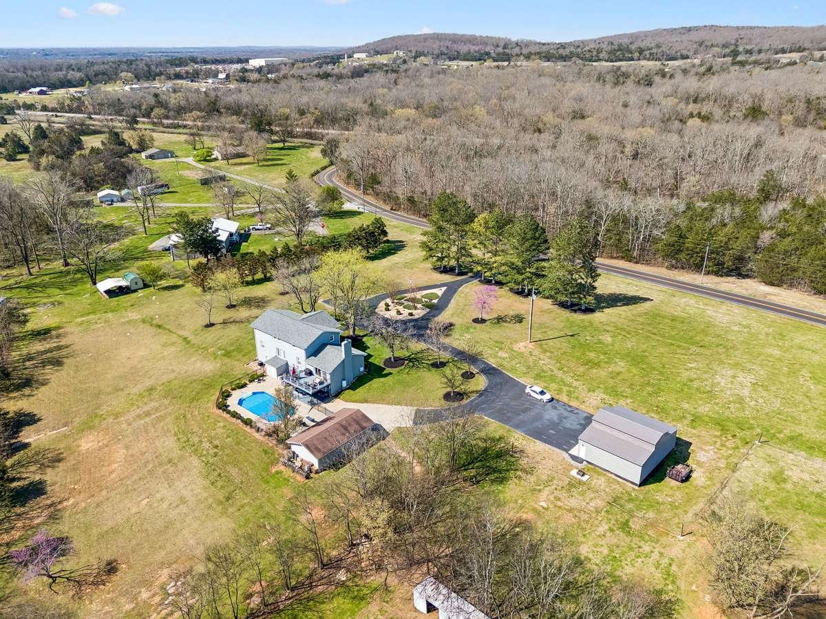 This sweeping aerial perspective showcases the complete 17-acre property with its wooded privacy, inground pool, detached garage, and three-bay shop positioned perfectly for both recreation and practical storage needs.