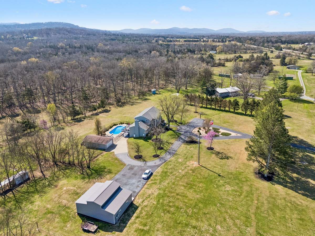 This aerial perspective showcases the property's thoughtful layout with the main home, detached garage, and three-bay workshop strategically positioned across the open pasture areas while maintaining the natural wooded buffer for privacy.