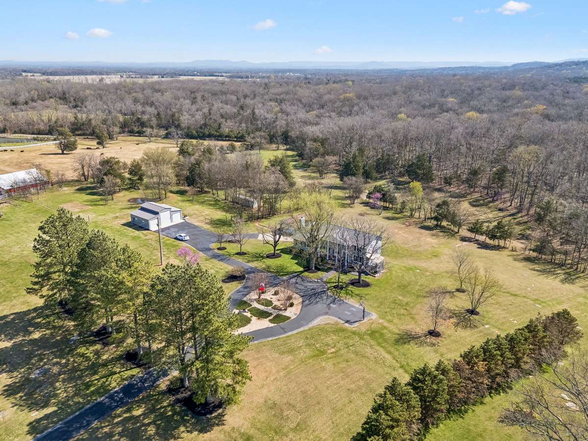 Aerial view showcases the colonial home positioned on its expansive wooded acreage with the detached garage and three-bay shop strategically placed for easy access while maintaining the privacy of the main house.