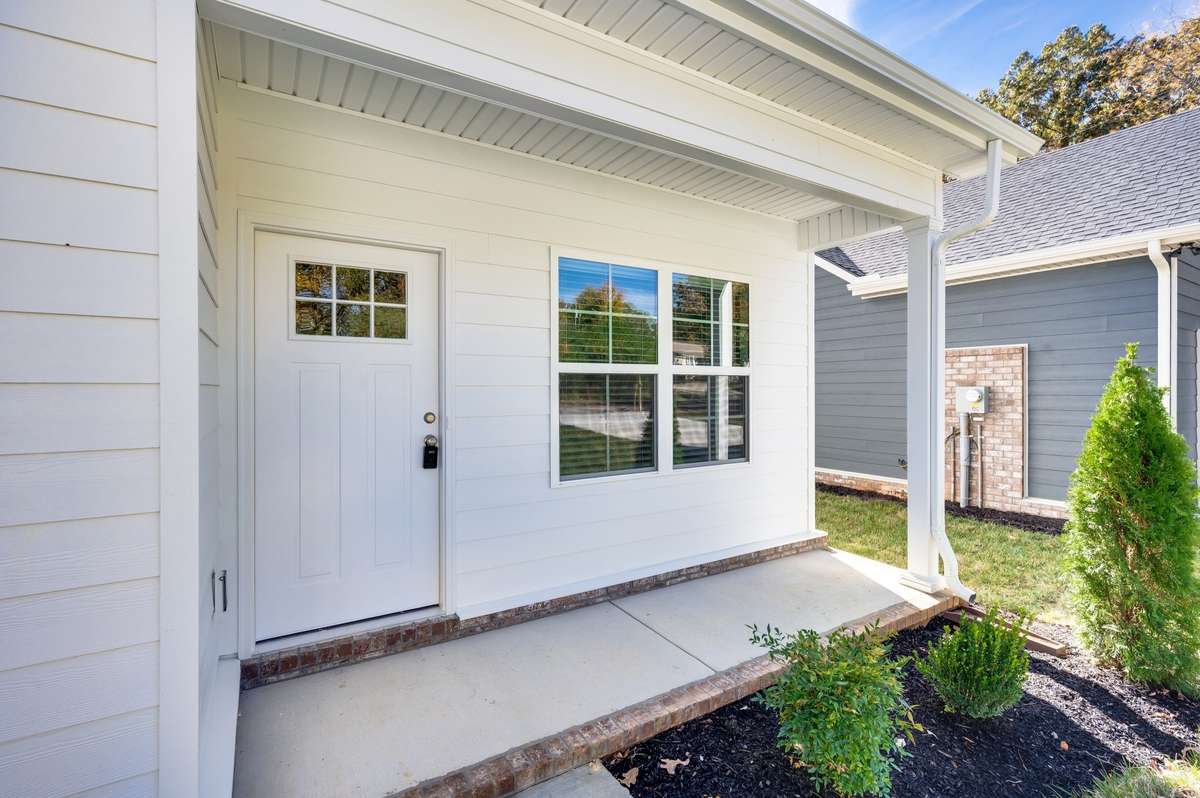 Welcoming covered entry with a modern front door and clean white siding. Brick trim and simple landscaping add charm, while the porch offers a cozy spot to greet guests or enjoy a quiet moment outdoors. Stylish, low-maintenance, and inviting.
