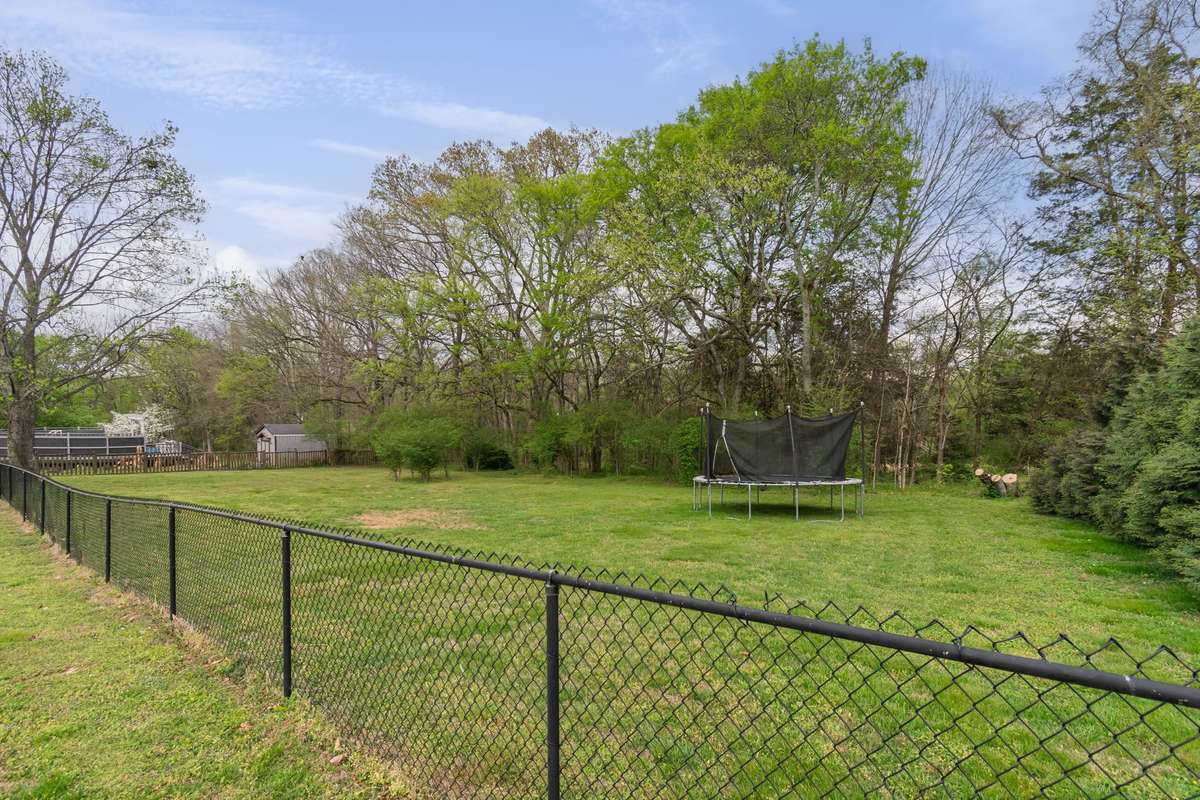 The private, fenced backyard is framed by mature trees and offers space for a trampoline, play set, or outdoor games. The yard extends past the wire fence into the tree line.