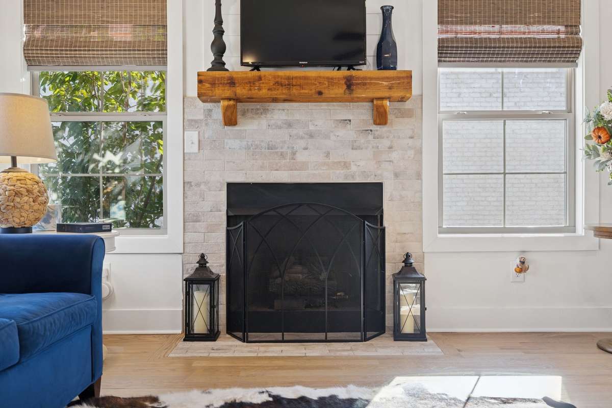 The gas fireplace features a charming brick surround, floor-level hearth, and classic wooden mantel. Large windows with substantial trim highlight the timeless character of the Craftsman style.