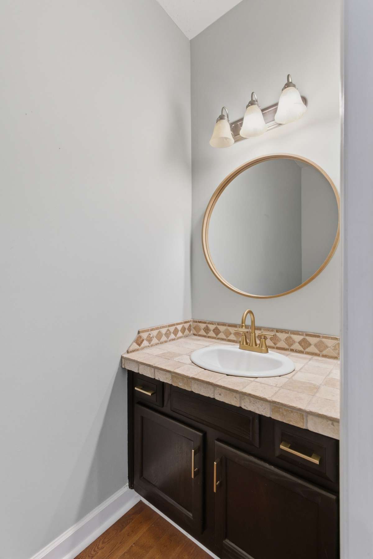Stylish main-level half bath featuring a tiled vanity countertop, brass fixtures, and a modern round mirror—perfectly positioned for guests and everyday convenience.