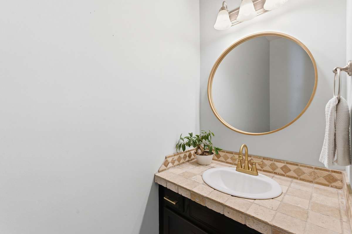 Stylish main-level half bath featuring a tiled vanity countertop, brass fixtures, and a modern round mirror—perfectly positioned for guests and everyday convenience.