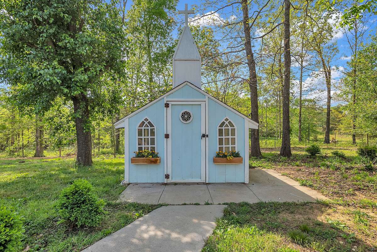 Adorable light-blue chicken coop with steeple, arched windows, and flower boxes sits tucked among the trees. Fully functional with easy access and a peaceful setting — the perfect setup for fresh eggs
