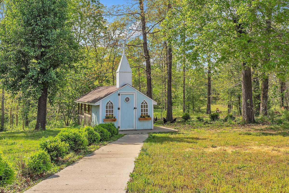 Unique chicken coop designed like a quaint blue chapel with steeple, arched windows, and blooming flower boxes. Ready for your flock with convenient access and a scenic wooded backdrop